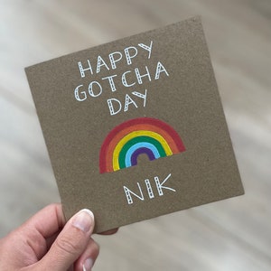 May include: A brown card with the text "Happy Gotcha Day" in white cursive writing. A rainbow with red, orange, yellow, green, blue, indigo, and violet stripes is drawn below the text. The name "Nik" is written in white cursive writing below the rainbow.