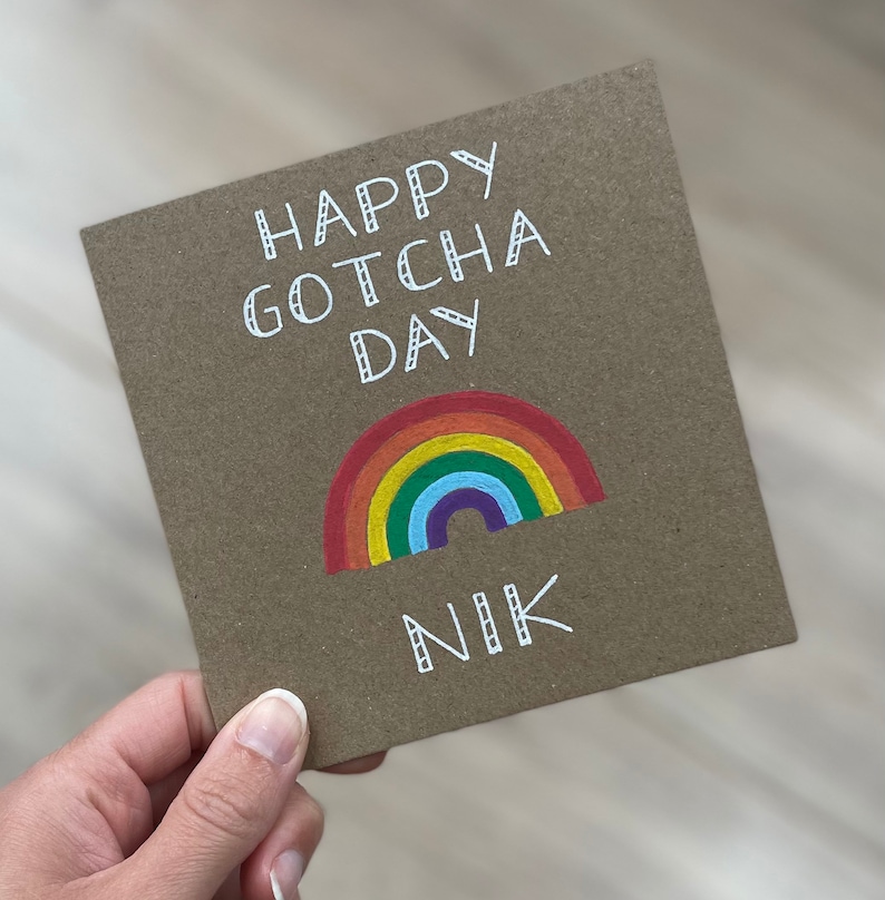 May include: A brown card with the text "Happy Gotcha Day" in white cursive writing. A rainbow with red, orange, yellow, green, blue, indigo, and violet stripes is drawn below the text. The name "Nik" is written in white cursive writing below the rainbow.