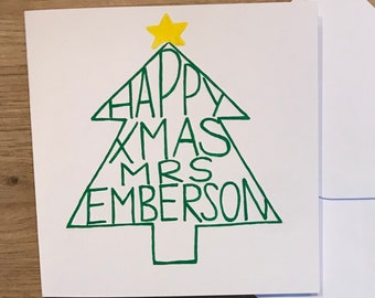 May include: A white Christmas card with a green Christmas tree design. The tree is filled with the words "HAPPY XMAS MRS EMBERSON". A yellow star is at the top of the tree. A white envelope is next to the card.