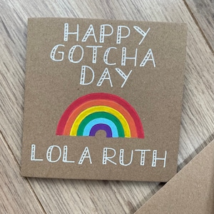 May include: A brown card with the text "Happy Gotcha Day Lola Ruth" in white outlined letters. A rainbow is drawn below the text.