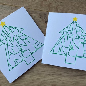 May include: Two white Christmas cards with a green Christmas tree design. The tree shape contains the words "HAPPY XMAS AUNT & UNCLE" in green. A yellow star is at the top of each tree. The cards are on a light brown wooden surface.