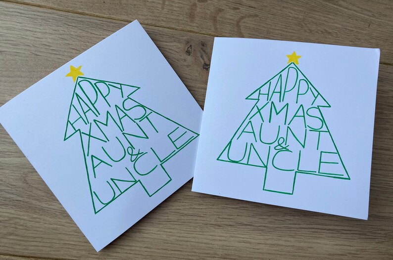 May include: Two white Christmas cards with a green Christmas tree design. The tree shape contains the words "HAPPY XMAS AUNT & UNCLE" in green. A yellow star is at the top of each tree. The cards are on a light brown wooden surface.