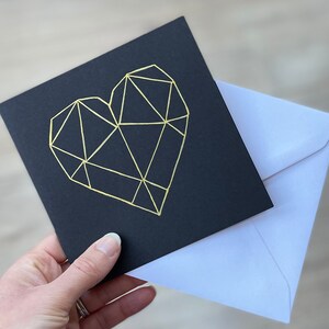 May include: A black card with a gold geometric heart design. The card is accompanied by a white envelope.