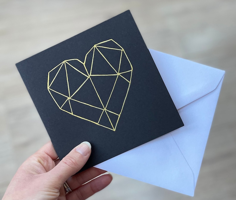 May include: A black card with a gold geometric heart design. The card is accompanied by a white envelope.