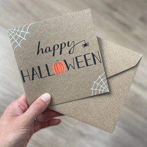 May include: A brown Halloween greeting card with the words "happy HALLOWEEN" in black lettering, a small orange pumpkin, and a black spider. White spiderwebs are in the corners. The card is accompanied by a matching envelope.