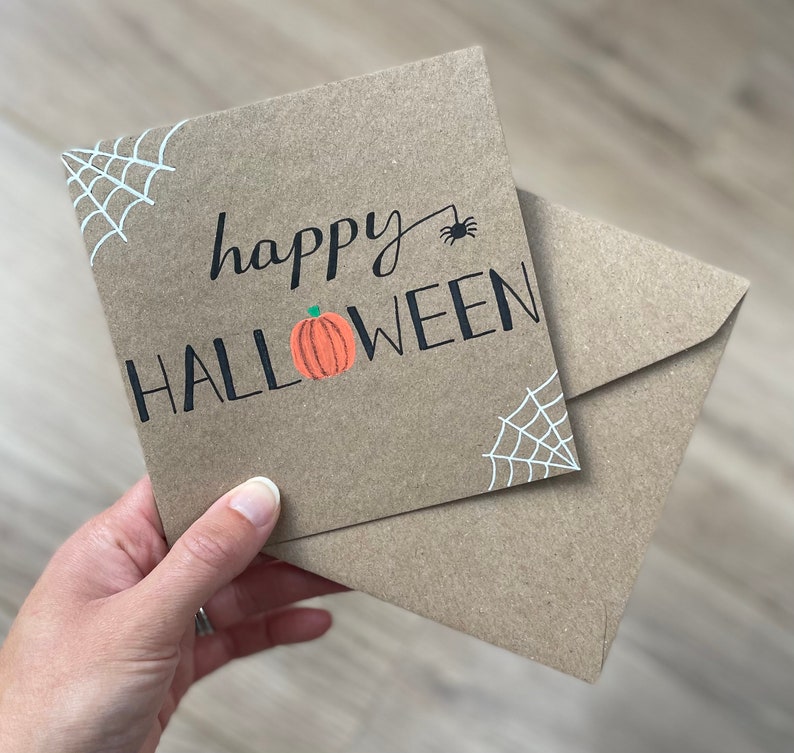 May include: A brown Halloween greeting card with the words "happy HALLOWEEN" in black lettering, a small orange pumpkin, and a black spider. White spiderwebs are in the corners. The card is accompanied by a matching envelope.