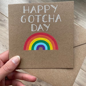 May include: A brown card with the text "Happy Gotcha Day" in white lettering. A rainbow with red, orange, yellow, green, blue, indigo, and violet stripes is drawn below the text.