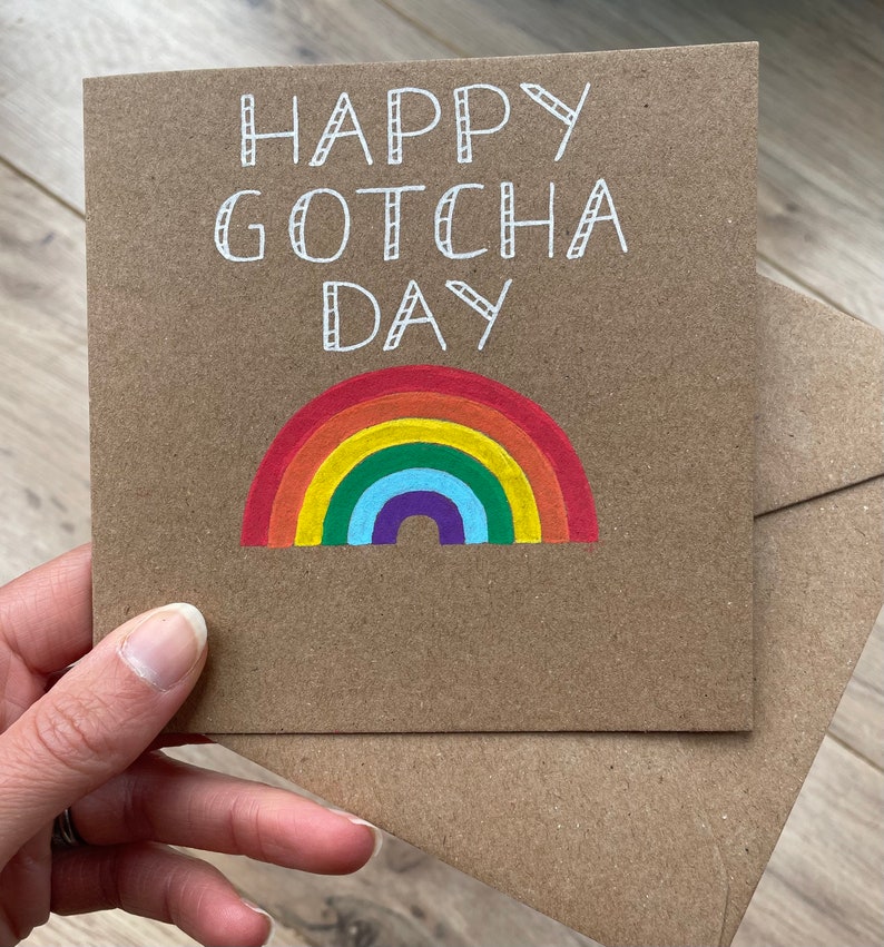 May include: A brown card with the text "Happy Gotcha Day" in white lettering. A rainbow with red, orange, yellow, green, blue, indigo, and violet stripes is drawn below the text.