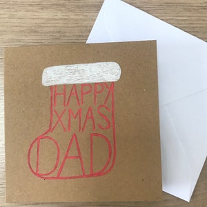 May include: A square, brown Christmas card with a red outline of a stocking that says "HAPPY XMAS DAD". The top of the stocking is white. A white envelope is next to the card.