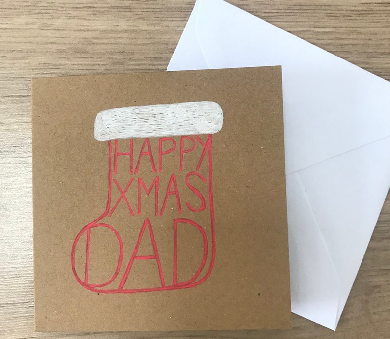 May include: A square, brown Christmas card with a red outline of a stocking that says "HAPPY XMAS DAD". The top of the stocking is white. A white envelope is next to the card.
