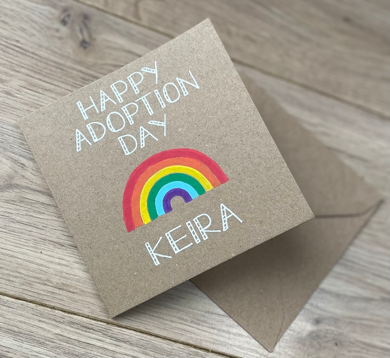 May include: A brown card with the text "Happy Adoption Day" and a rainbow with the name "Keira" written below it.