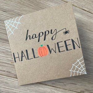 May include: A square, brown card with the words "happy HALLOWEEN" in black script and block letters. A small orange pumpkin with a green stem is between the words. White spiderwebs are in the corners, and a black spider is above the word "Halloween".