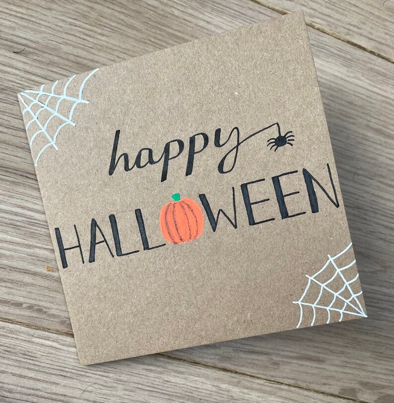 May include: A square, brown card with the words "happy HALLOWEEN" in black script and block letters. A small orange pumpkin with a green stem is between the words. White spiderwebs are in the corners, and a black spider is above the word "Halloween".