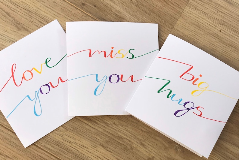 May include: Three white cards with rainbow colored lettering. The first card says "love you", the second card says "miss you", and the third card says "big hugs".