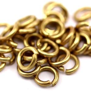 Könnte beinhalten: Nahaufnahme eines Haufens kleiner, offener Sprungringe in einem warmen, messingfarbenen Goldton. Die Ringe sind vor einem weißen Hintergrund verstreut, wobei sich einige Ringe überlappen. Diese Schmuckzubehörteile werden zum Basteln und zur Schmuckherstellung verwendet.