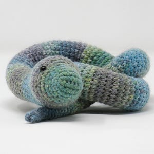 May include: A handmade crochet snake toy in shades of blue, green, and grey. The snake is curled up, with a rounded head and small, attached limbs. The toy is made of soft yarn.