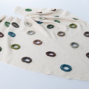 May include: Cream-colored knitted blanket with multiple colorful ring-shaped embellishments. The rings are in shades of brown, green, and blue, adding a playful contrast to the neutral background. The blanket is folded and draped, showcasing its texture.