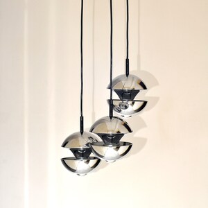Peut inclure: Trois lampes suspendues chrom&eacute;es avec un design sph&eacute;rique en couches pendent &agrave; des cordons noirs contre un mur blanc.