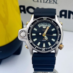 Può includere: Un orologio Citizen Eco-Drive argento e nero con un cinturino in gomma nera. Il quadrante dell'orologio ha uno sfondo nero con marcature bianche e un display digitale. L'orologio ha una ghiera dorata con un anello rotante che arriva fino a 50. L'orologio è su un supporto con la parola "Raymond" su di esso.