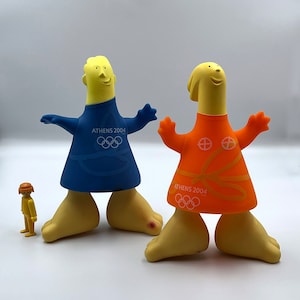 Può includere: Due figure di cartoni animati gialle e arancioni con il testo "ATHENS 2004" e gli anelli olimpici sulle loro maglie. Le figure sono in piedi su una superficie bianca.