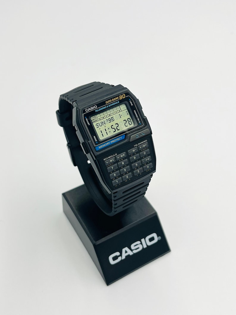 Op de afbeelding: Een zwart Casio Data Bank 80 rekenmachinehorloge met een digitaal display dat de datum, tijd en dag van de week weergeeft. Het horloge heeft een zwarte resin band en een zwart rekenmachinetoetsenbord.