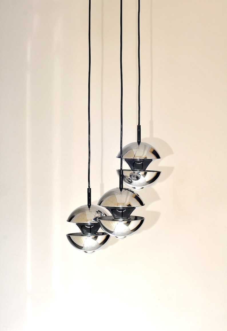 Peut inclure: Trois lampes suspendues chrom&eacute;es avec un design sph&eacute;rique en couches pendent &agrave; des cordons noirs contre un mur blanc.