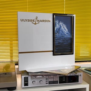 May include: A white display featuring the gold text "ULYSSE NARDIN" and an anchor logo. A framed picture of a blue wave is on the right. The display is on a stereo receiver with a record player and a vintage radio.