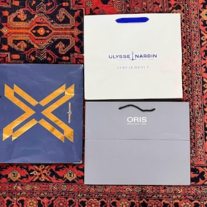 May include: Four shopping bags on a patterned rug. The bags are white, blue, and gray, with logos for Ulysse Nardin and Oris. The Ulysse Nardin bag has a blue handle and the text "#FREAKMEOUT". The Oris bag has a black handle.