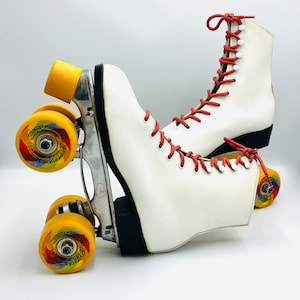 以下が含まれることがあります： 黄色い車輪と赤い靴ひもが付いた白いローラースケート。車輪は虹のデザインで、「FREE RIDE」の文字が書かれています。