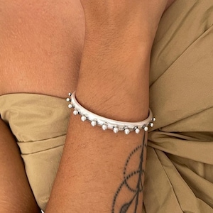 May include: A silver-toned chain link necklace, a silver-toned bangle bracelet with small spherical studs, and two silver-toned rings featuring similar spherical stud detailing. The jewellery pieces are metallic with a polished finish.