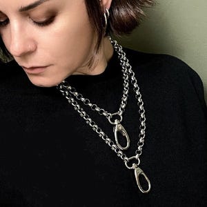 Pode incluir: Um colar de corrente prateada com dois fechos lagosta integrados, usado sobre uma camisa preta. O colar consiste em elos grossos e arredondados e inclui um conjunto de pulseiras prateadas a condizer no pulso.