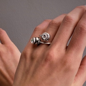 Könnte beinhalten: Ein silberfarbenes Armband mit einem dreieckigen Mittelstück und einem dunklen Stein, kombiniert mit zwei silberfarbenen offenen Ringen, die jeweils mit einem einzelnen klaren runden Stein besetzt sind, getragen an Handgelenken und Fingern.