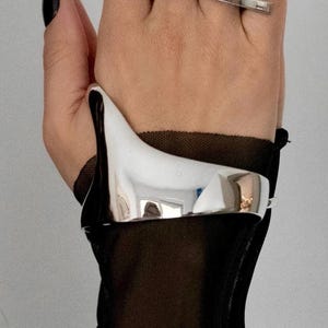 May include: A silver ring with a pointed design and a wide silver cuff bracelet. The jewelry is worn on a hand with black nail polish. The background is a plain white wall.