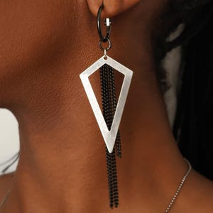 Op de afbeelding: Geometrische zilveren oorbellen met zwarte kettingfranjes. De oorbellen hebben een ruitvormig ontwerp met een geborstelde metalen afwerking en een zwarte ring. Een modern accessoire.