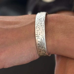 May include: A silver-toned cuff bracelet and two matching rings, each featuring an intricate, embossed floral pattern. The jewellery pieces have a polished metallic finish with detailed scrollwork engravings across the surface of the metal.