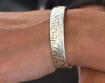 May include: A silver-toned cuff bracelet and two matching rings, each featuring an intricate, embossed floral pattern. The jewelry pieces have a polished metallic finish with detailed scrollwork engravings across the surface of the metal.