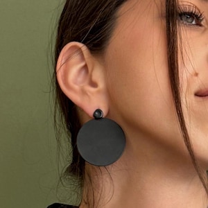 Puede incluir: Pendientes redondos negros mate con un pequeño detalle de tachuela. Los pendientes tienen un diseño sencillo y moderno. Los pendientes son llevados por una persona.