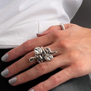 May include: A silver-toned chain link bracelet featuring a central clear crystal stone and a matching silver-toned ring with two clear crystal stones. The jewellery pieces have a polished, metallic finish and a chunky, interconnected link design.