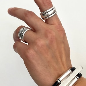 May include: Two polished silver rings with a triple-band design worn on the index and ring fingers, paired with a black leather cord bracelet featuring a central silver bar and two cylindrical silver accents on a white background.