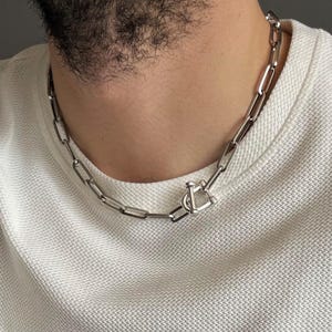 Könnte beinhalten: Eine silberfarbene Kette mit rechteckigen Gliedern und einem T-Bar-Verschluss. Die Person trägt ein weißes, strukturiertes Strickoberteil. Ein silbernes Armband ist am Handgelenk zu sehen. Ein stilvolles Accessoire.