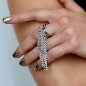 May include: A silver-toned metal ring featuring a cluster of delicate hanging chains and a matching wide cuff bracelet with a circular toggle closure. The jewellery set is crafted from polished metal with a smooth, reflective finish on the cuff.