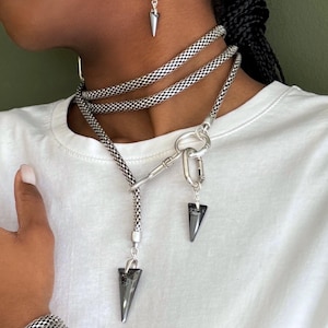 May include: A collection of silver-toned jewelry, including a necklace, earrings, bracelet, and ring. The necklace features a chain design with a pendant. The earrings have an oval shape with a spike. The bracelet and ring are also silver.