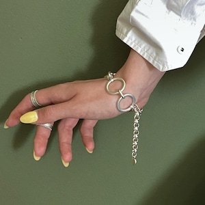 Può includere: Un bracciale a catena color argento con due prominenti maglie circolari come punto focale, indossato al polso insieme a due sottili anelli color argento. Il bracciale include un'estensione a catena pendente ed è abbinato a una camicia bianca.