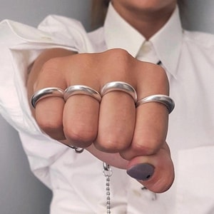 Könnte beinhalten: Silberne Knöchelringe an vier Fingern. Die Ringe sind dick und abgerundet, mit einer glatten, polierten Oberfläche. Die geballte Hand zeigt die Ringe vor einem weißen Hemd.