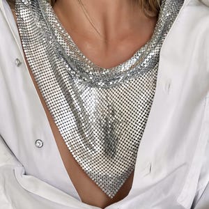 May include: A silver mesh necklace with a draped design. The necklace is worn over a white button-down shirt. The model is also wearing silver earrings and rings. The jewelry is a statement piece.