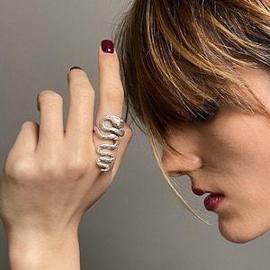 May include: A silver-toned ring shaped like a coiled snake with a textured surface, worn on a finger. The ring features a detailed, winding body design that wraps around the finger, crafted from a metallic material with a polished finish.