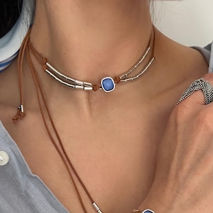 May include: A brown leather cord choker and matching bracelet set, each featuring a central square blue stone framed in silver, flanked by silver cylindrical beads. The choker has long dangling ends with silver tips, and the bracelet is adjustable.