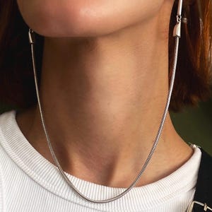 May include: A silver-toned earring featuring a long, thin vertical bar attached to a flexible snake chain that loops under the chin. The chain connects back to the bar, creating a continuous, draped effect that hangs from the earlobe.