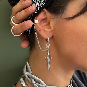 Op de afbeelding: Een zilverkleurige oormanchet verbonden door een fijn kettinkje aan een oorring met een kleine zwaardhanger. Het sieraad is gemaakt van gepolijst metaal en bestaat uit een eenvoudige oorring en een gladde oormanchet voor het bovenste kraakbeen.