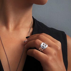 Könnte beinhalten: Eine lange, dünne Kugelkette aus dunklem Metall mit einem einzelnen, glatten, schwarzen, kegelförmigen Anhänger. Dazu wird ein breiter, polierter Ring in Silberoptik getragen, der ein ineinandergreifendes, abgerundetes Gliederkettendesign aufweist.